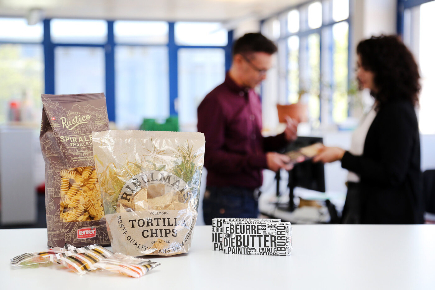 Au premier plan, des prototypes de sachets, sachets et films d'emballage pour le beurre. En arrière-plan, deux personnes en train de discuter. (