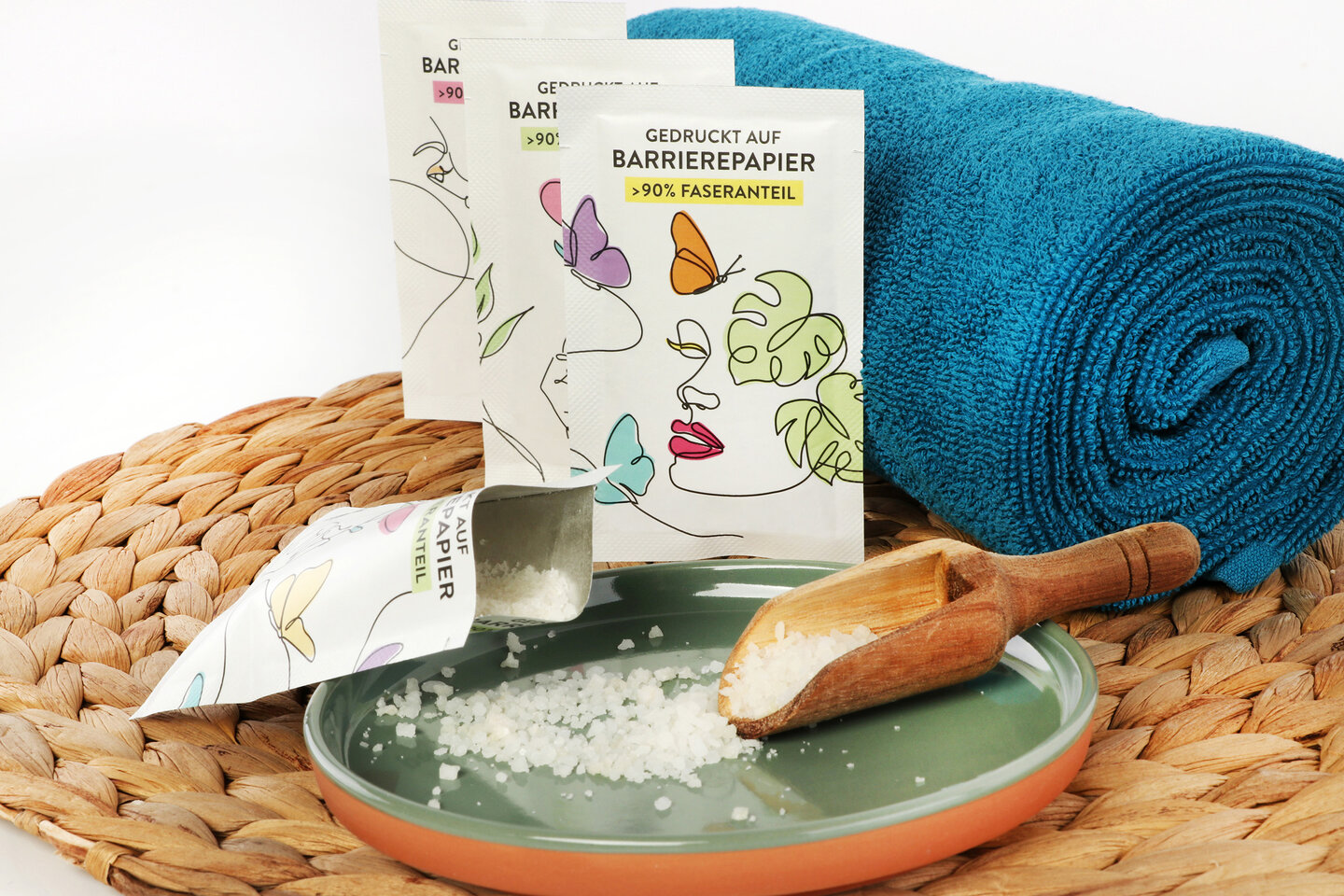Trois emballages en papier barrière contenant des sels de bain sont appuyés contre un rouleau de serviettes.