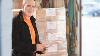 Un homme se tient devant une palette de cartons emballés sous film étirable.