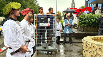 Deux hommes dans un chariot grillagé, entourés d'employés, attendent le baptême de l'imprimante.