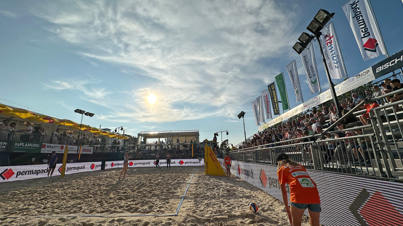 Terrain central de beach-volley dans le soleil couchant. La bande publicitaire affiche le logo Permapack.