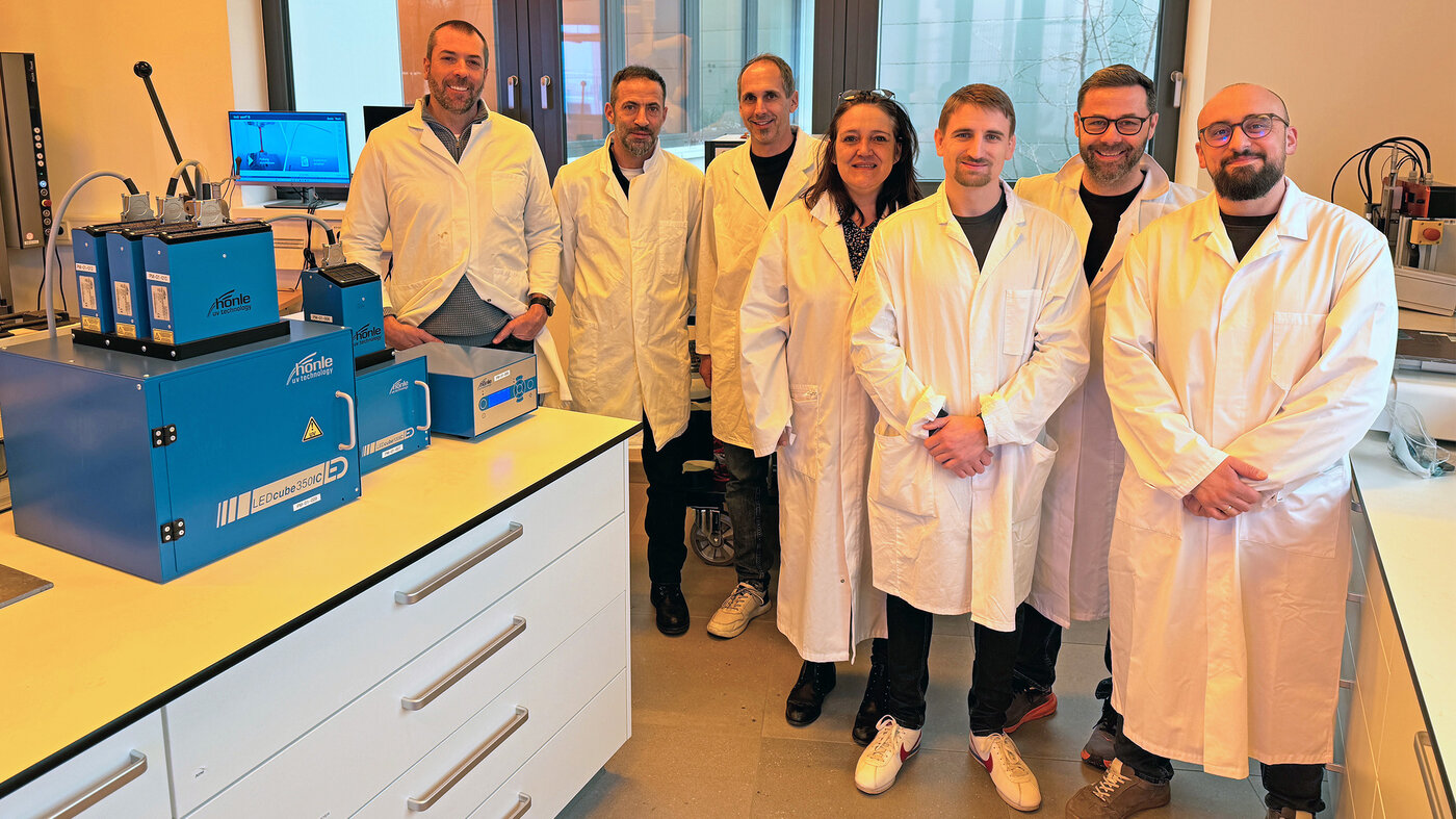 Groupe d’employés en blouses de laboratoire. Au premier plan, des appareils de durcissement aux UV et des appareils de laboratoire pour les tests d’adhésifs.