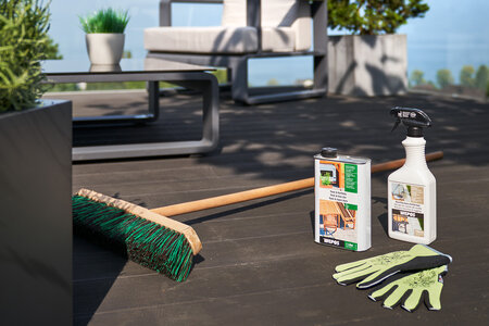 Terrasse en bois éclairée par le soleil, un balai deux produits de nettoyage et une paire de gants de jardinage disposés sur le sol